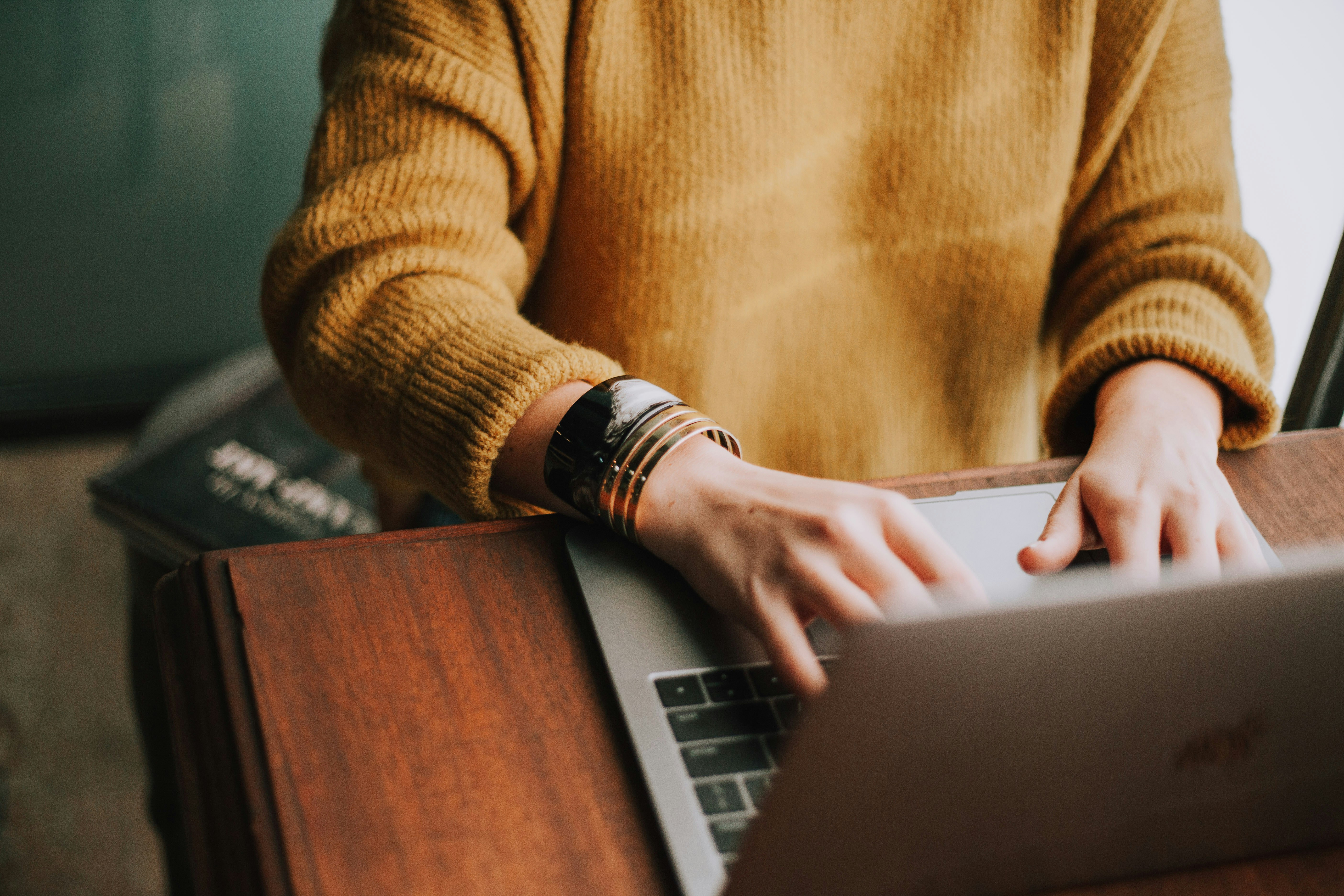 Hands typing on a laptop in a warm sweater, sunlit. A real workday, somewhere worth being.