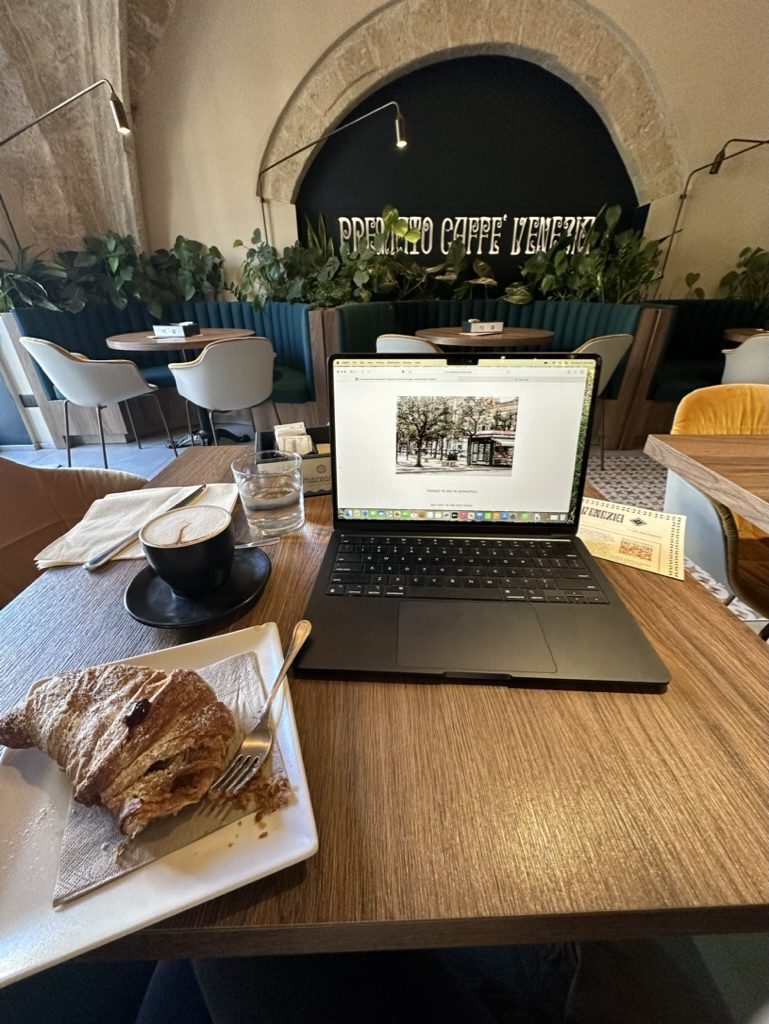 A laptop and latte on a cafe table by a sunlit window. A real workday, somewhere worth being.
